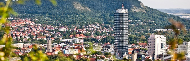 Blick auf eine Stadt mit einem markanten zylinderförmigen Gebäude und mit Bergen im Hintergrund