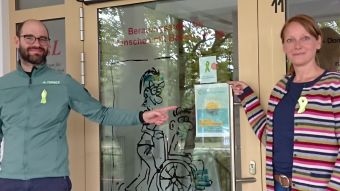 Ein Mann und eine Frau stehen vor einer Glastür, an die ein Rollstuhlfahrer mit Begleitperson gemalt ist, und tragen die Grüne Schleife, deren Symbol auch an der Tür hängt