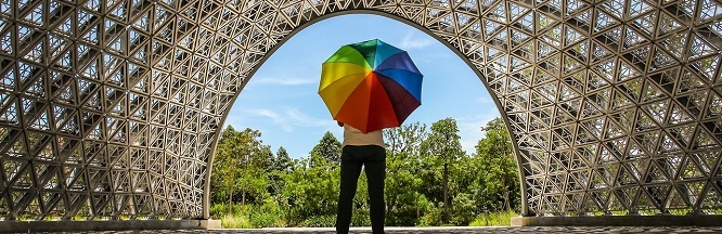 Person mit buntem Regenschirm steht unter bogenförmiger Metallkonstruktion