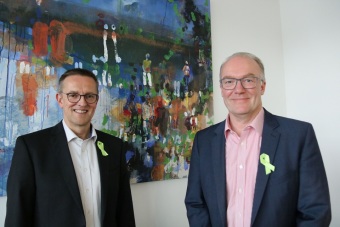 Zwei Männer stehen vor einer weißen Wand mit einem abstrakten Gemälde und tragen beide eine Grüne Schleife am Revers.