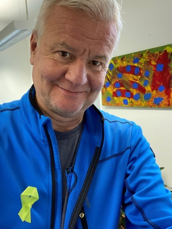 Ein Mann fotografiert sich im Büro selbst mit einer Grünen Schleife an der Jacke, im Hintergrund ist ein abstraktes Gemälde zu sehen